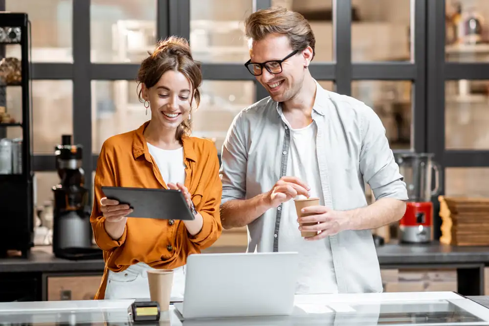 A woman holding a tablet showing the screen to a man who is holding a drink.