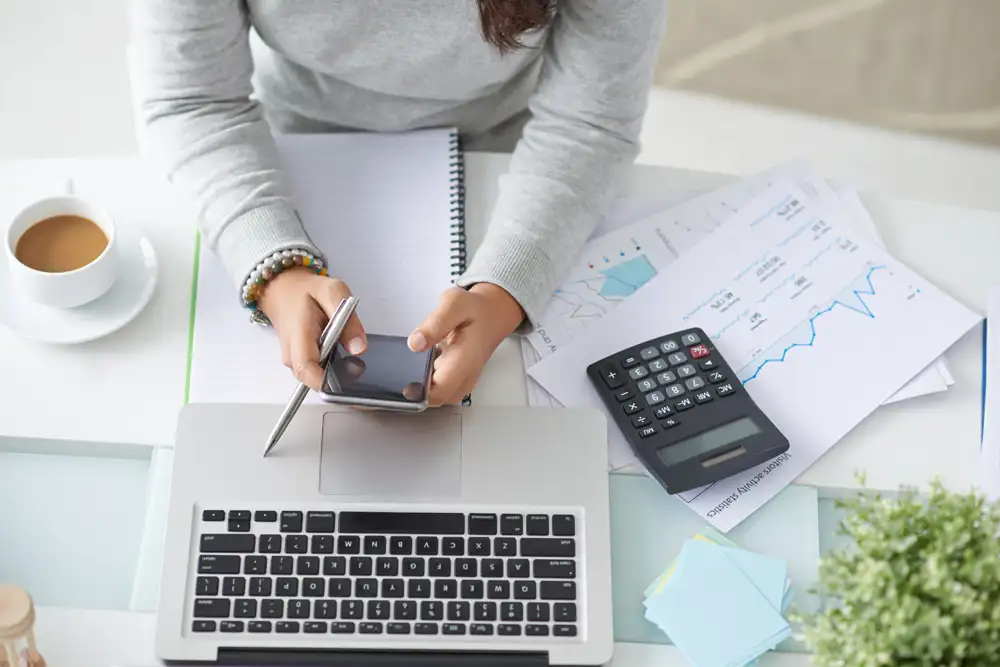 A woman's arm and hand using her phone next to her laptop and calculator.