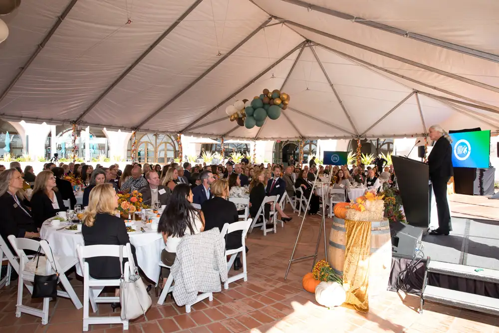 The CEO of Montecito Bank & Trust gives a speech to an audience of people sitting around large tables during an event.