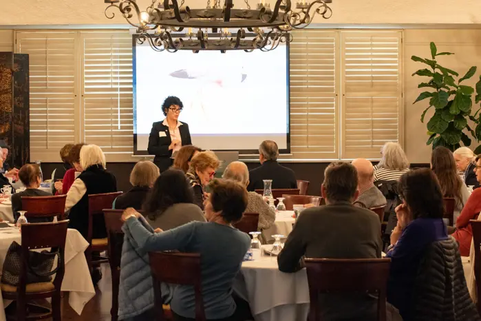 A bank employee gives a lecture to a group of elder adults sitting around large dinner tables.