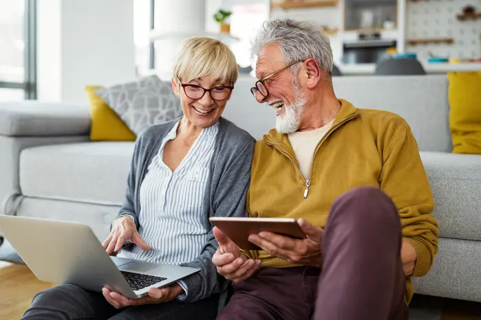 An elder couple lean against a couch smiling with a tablet in their hand.