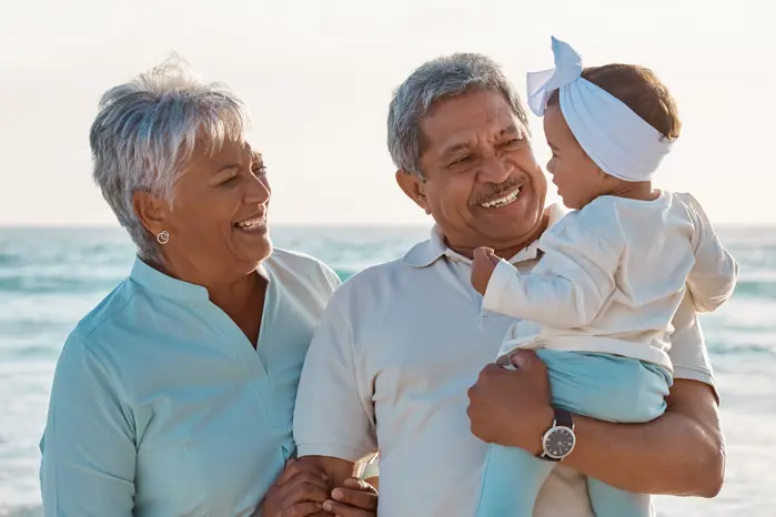 An elder couple are at the beach smiling and holding a baby.